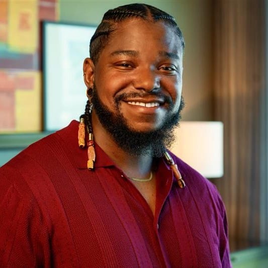 Smiling man in red shirt with braided hair.