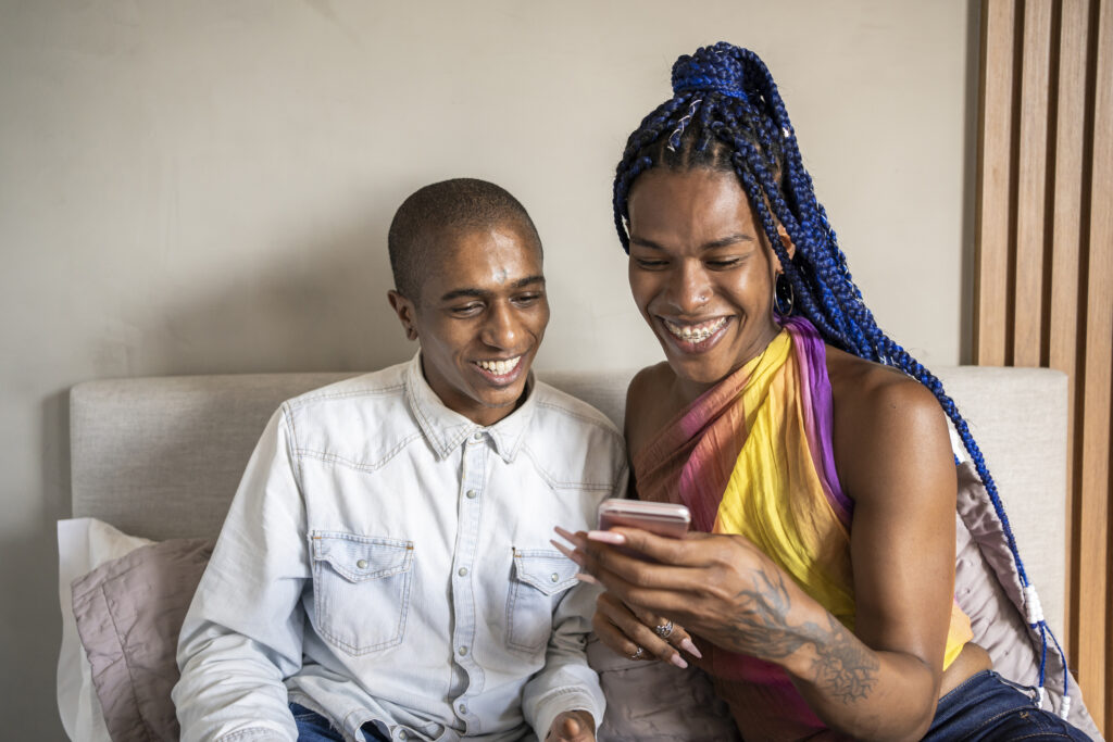 african american non binary person and transgender woman in bed using smart phone and talking. lgbt couple.