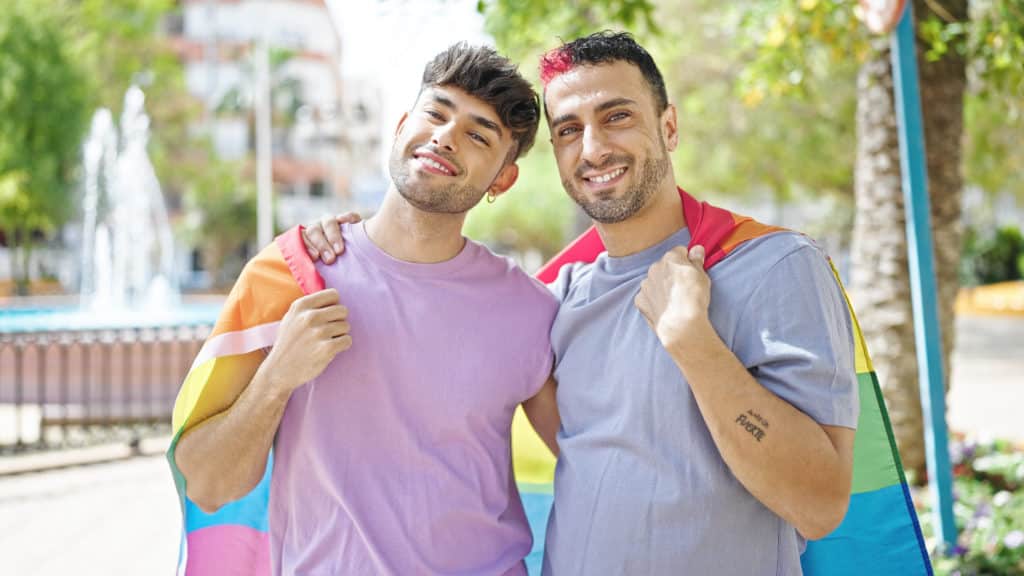 two men couple hugging each other holding homosexual flag at park